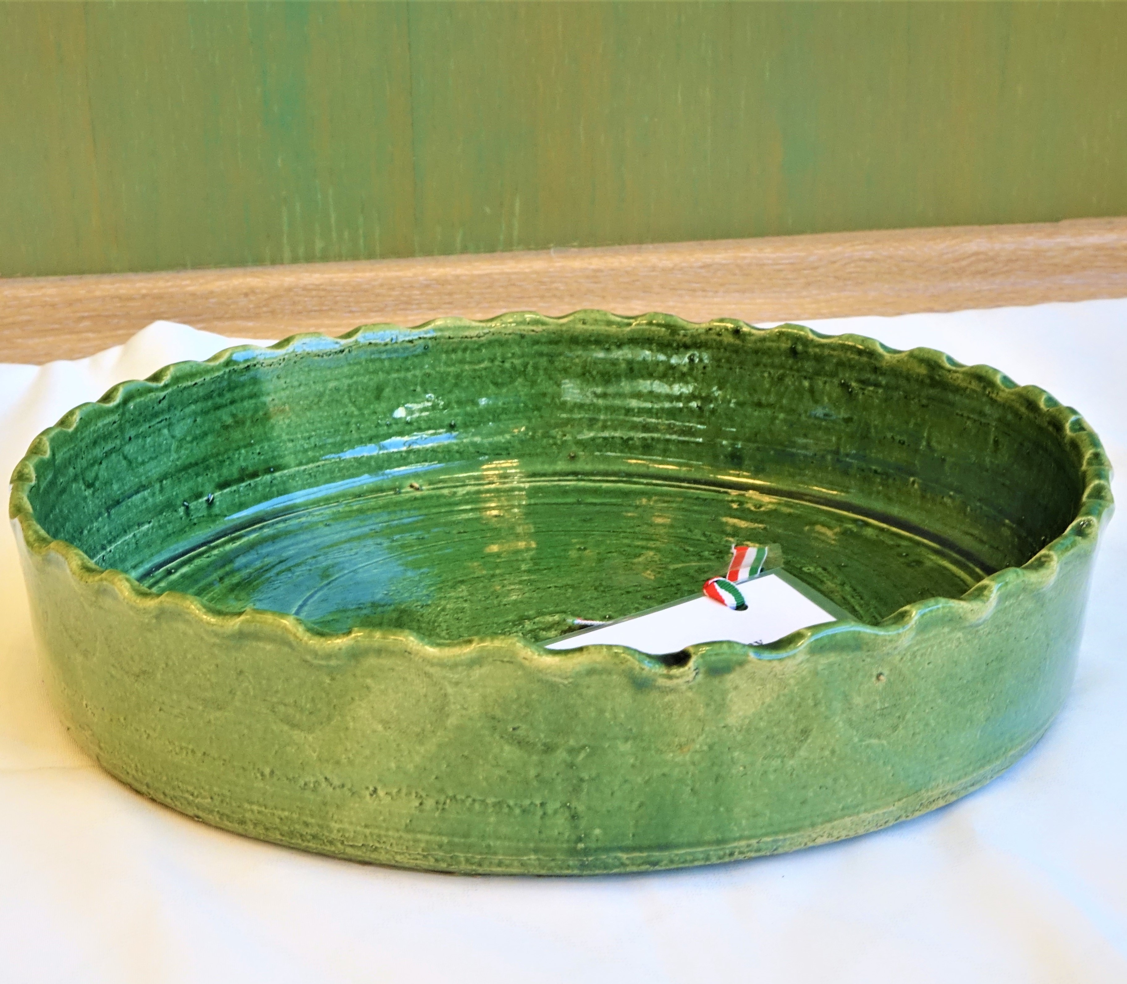 A képen egy kör alakú körülbelül harminc centiméteres kerámia pizzasütő tál látható néhány centiméteres fodros peremmel. Színe zöld, felszíne sima fényes.