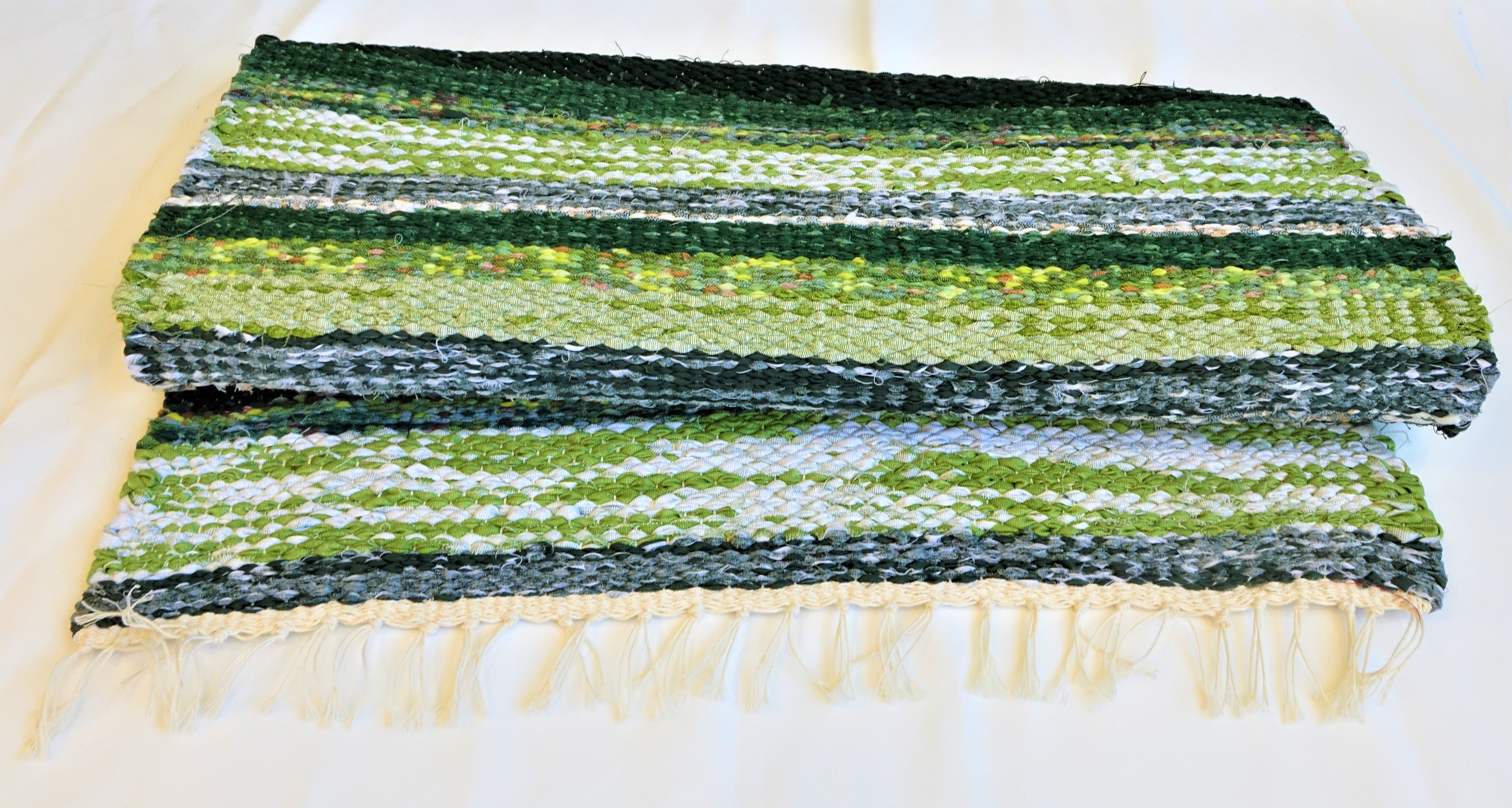 A képen egy körülbelül 1 méter hosszú és 70 centiméter széles, szőtt rongyszőnyeg látható. A szőnyeget keresztben többféle árnyalatú zöld, sárga, krémszínű, fehér és fekete csíkok mintázzák.