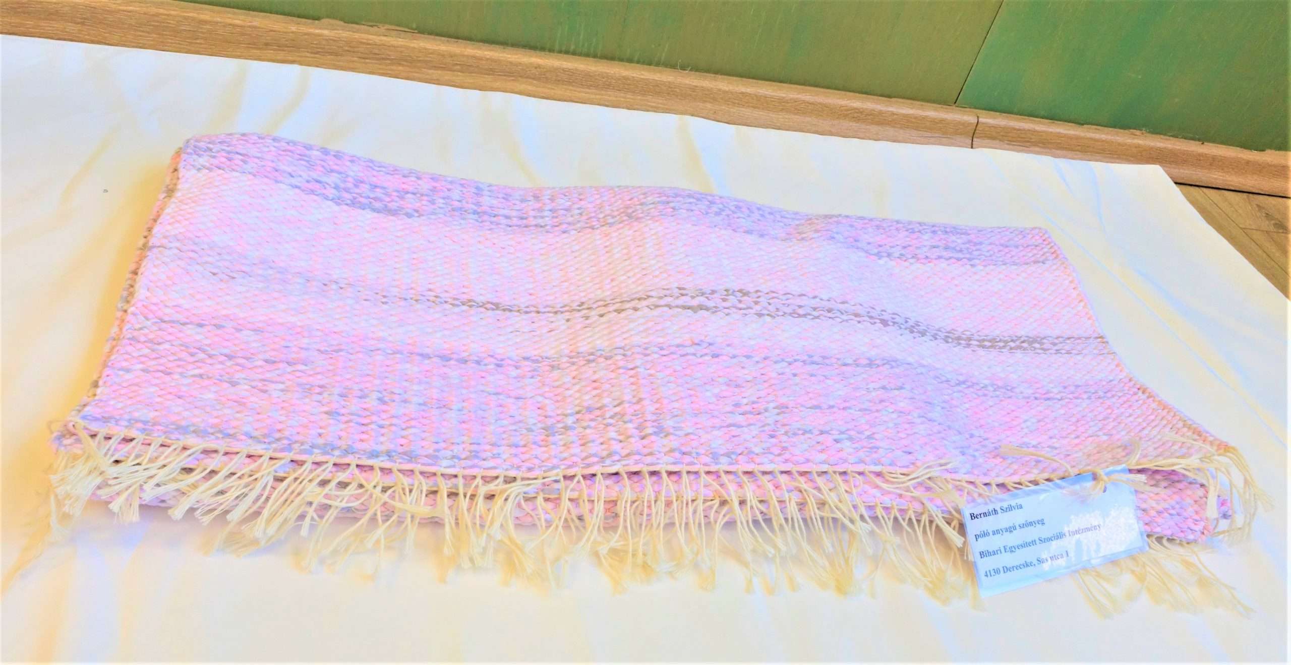 A képen egy póló alapú szőtt szőnyeg látható rózsaszín, szürke, fehér színben cirmos mintával. Végein rojtos. 