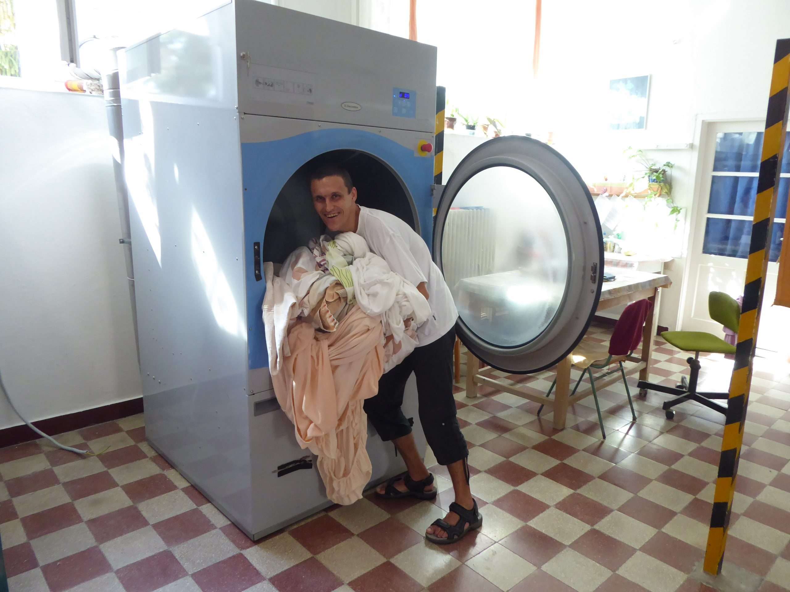 A fotón Deli István látható a mosodában, munkavégzés közben. A mosoda napfényes, középen egy nagy ipari mosógép látható szürke és kék színben. A mosógép ajtaja nyitva, a pályázó a mosott ruhákat szedi ki a gépből.