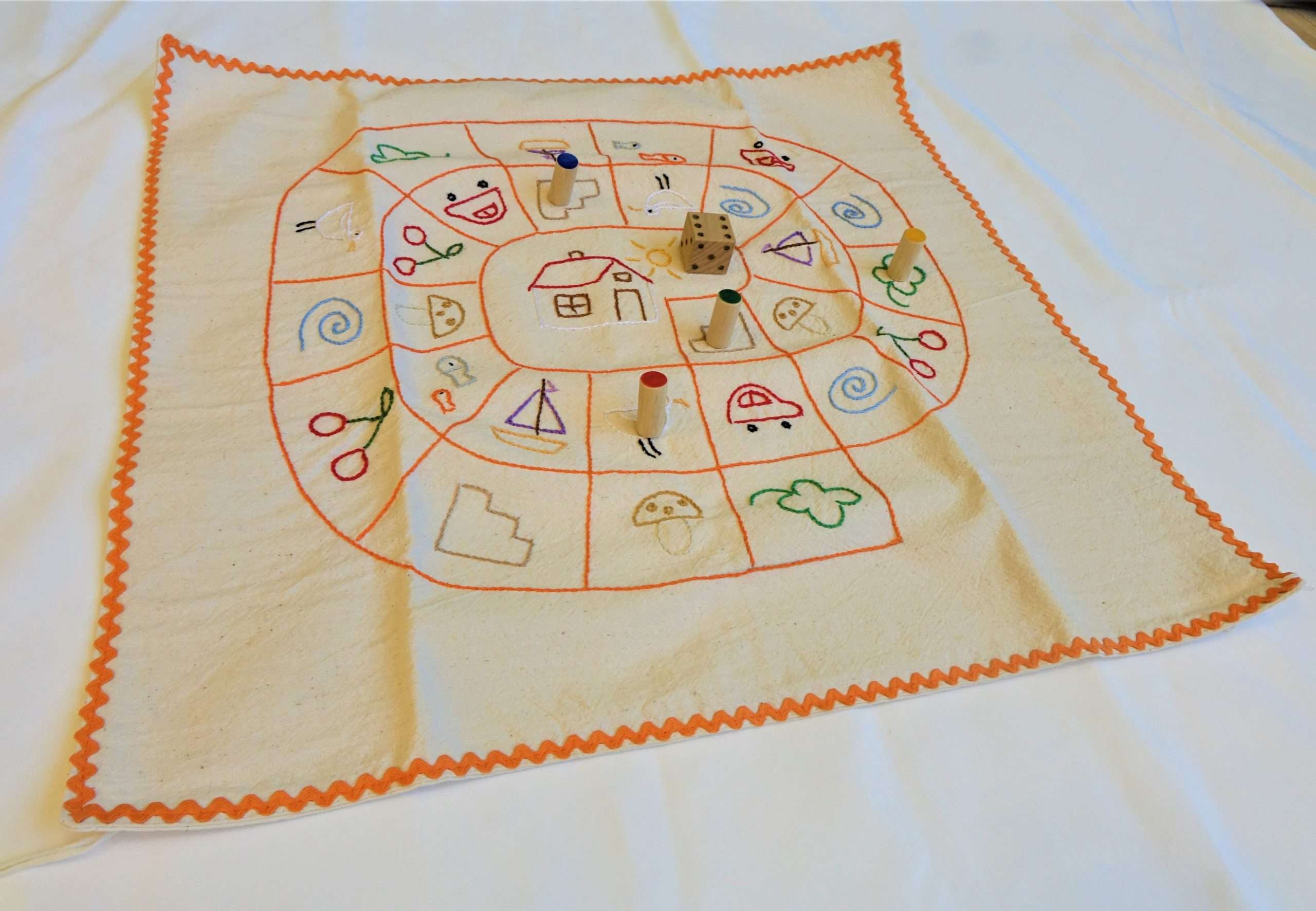 A képen egy kézzel készített textil társasjáték látható 4 bábuval és egy dobókockával. A natúr színű anyagon színes cérnával hímzett motívumok láthatóak, minden mező más motívumú. A társasjáték szélét narancssárga hullámos szegély díszíti.