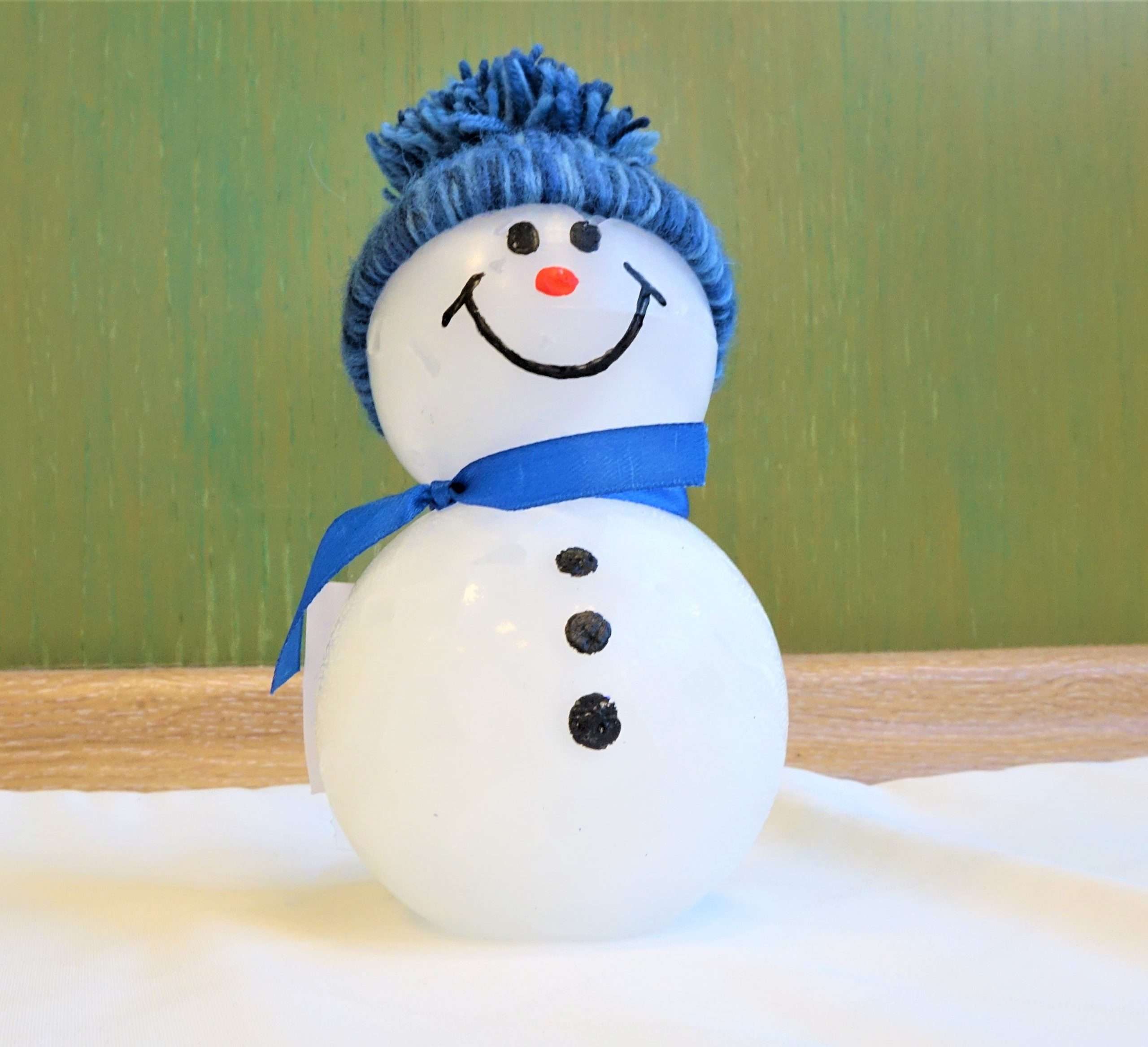 A képen egy körülbelül 15 centiméteres viaszból készült hóember látható. A hóember két szintből áll. Hasán 3 fekete gomb látható. Arca két szemből, egy narancssárga orrból és egy mosolygós szájból áll. Kék bolyhos fonott sapkát visel. Nyakában kék sál található szalagból.