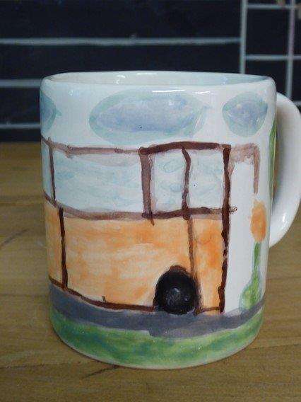 A fotón egy fehér csésze látható, melyre egy busz lett felfestve. A busz felett kék felhők láthatóak.