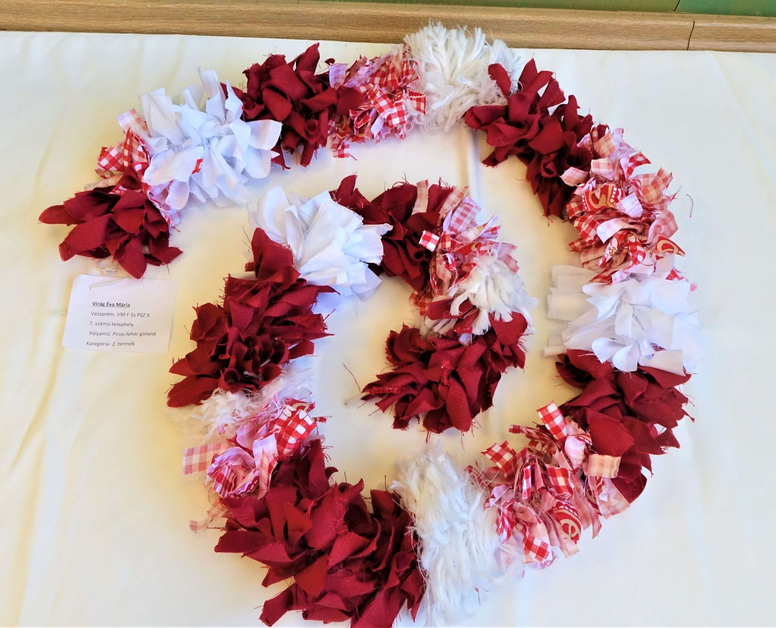 A képen egy körülbelül egy méter hosszú girland látható, amelyet piros és fehér textilcsíkokból készült virágok alkotnak. 